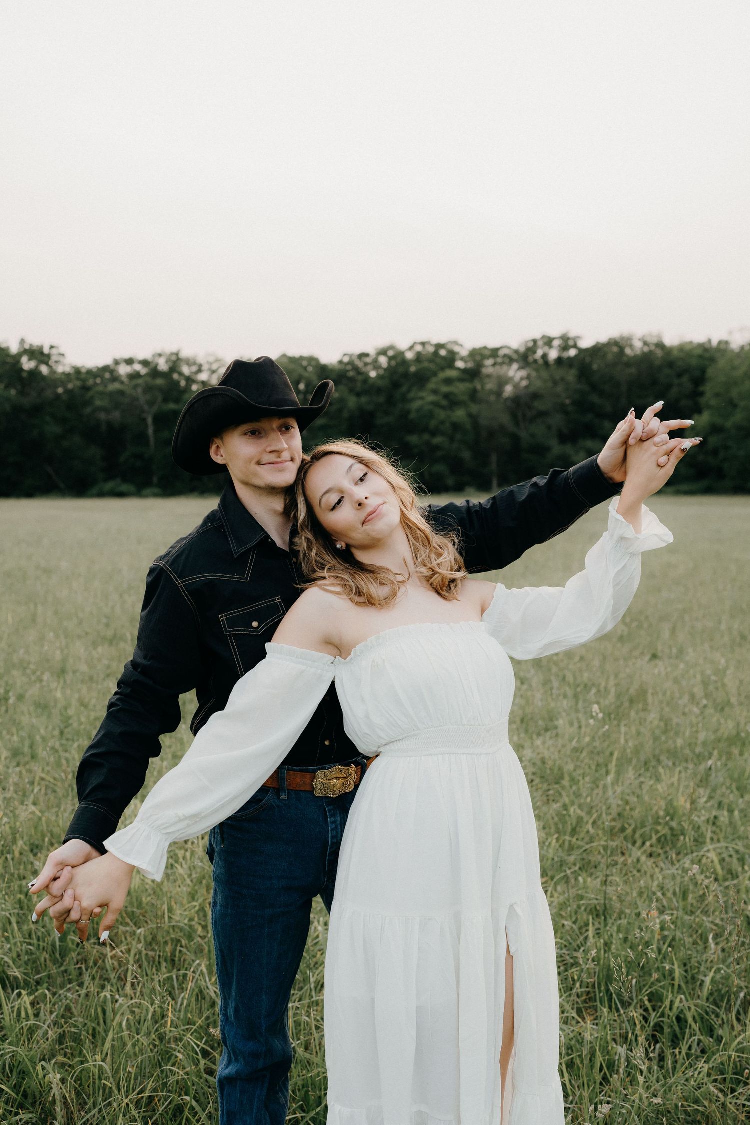 A joyful couple in western and wedding attire pose with outstretched arms in a field.