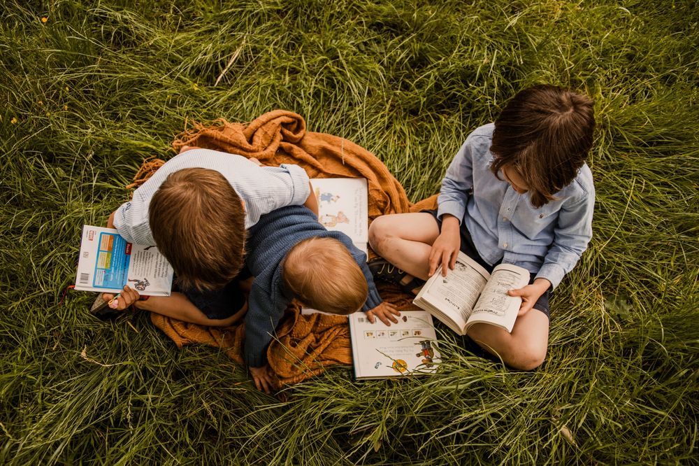 Three children sit together on an orange blanket in tall grass while reading and writing in journals outdoors.