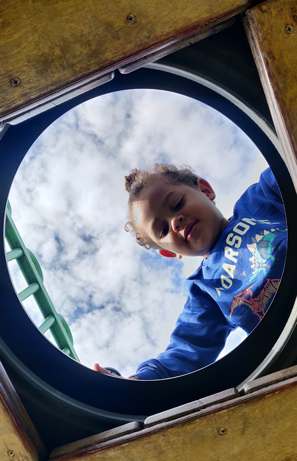 A boy looks down through a circular opening shows a blue sky with white puffy clouds beyond a wooden frame.