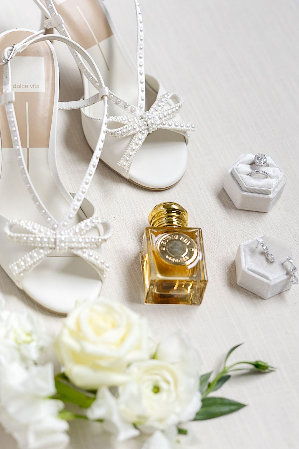 Elegant wedding accessories including white high heels, pearl necklace, perfume bottle and white roses on light background.