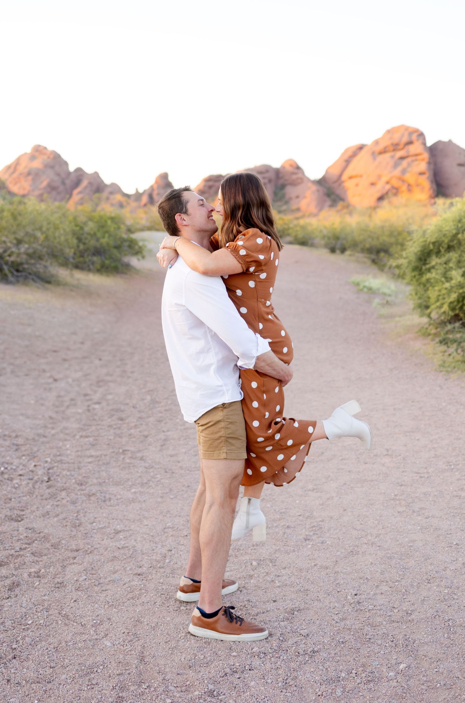 A couple embraces on a desert trail with red rocky mountains in the background during golden hour lighting.
