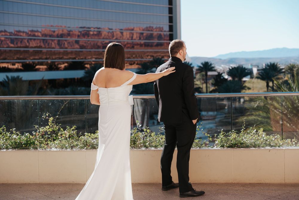 Two figures in formal wedding attire embrace while standing on an outdoor terrace with mountain views in the background.