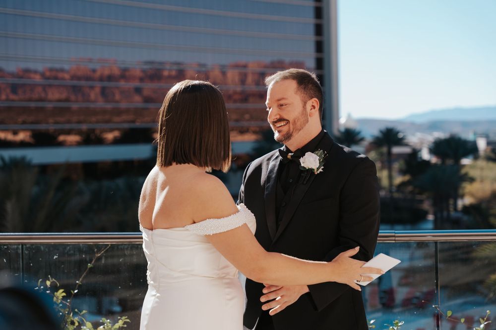 Two people share a joyful moment during their wedding ceremony on an outdoor balcony with city views.