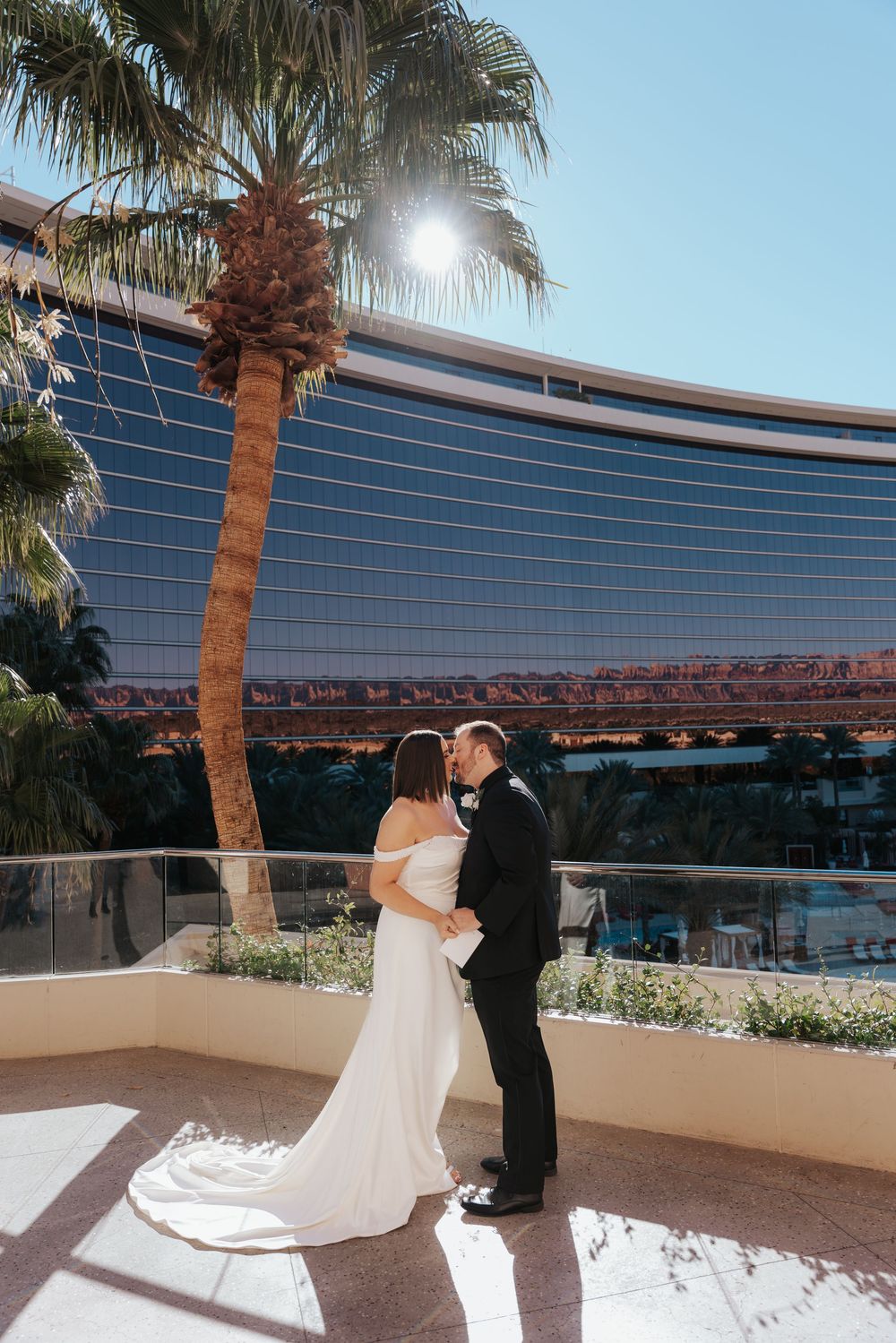 Wedding couple embraces on balcony of curved glass hotel building with palm trees in Las Vegas.