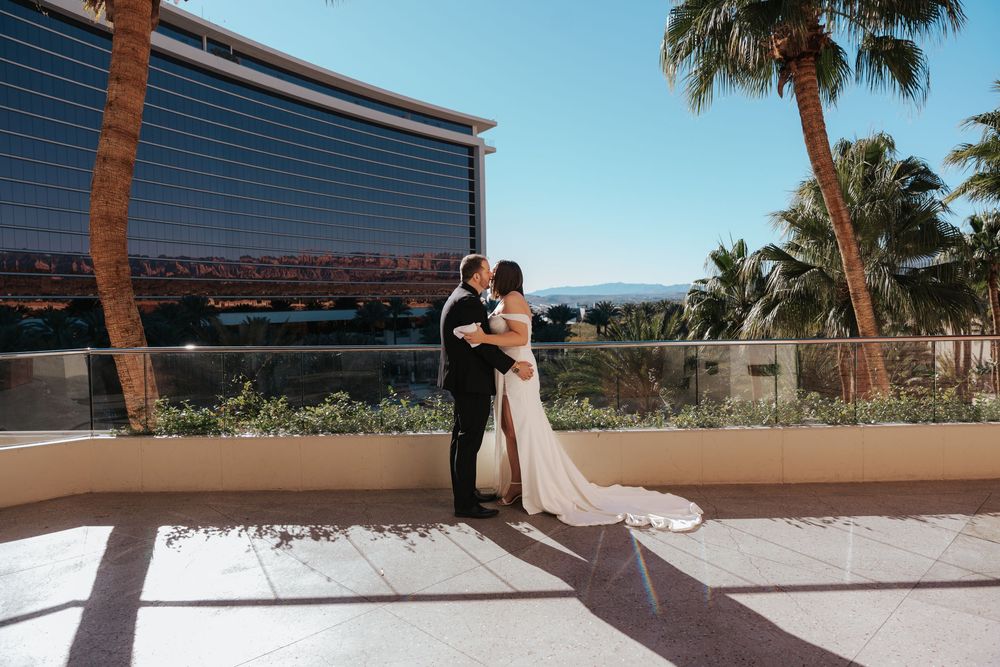 A couple in wedding attire embraces on a balcony with palm trees and a modern resort building in the background.