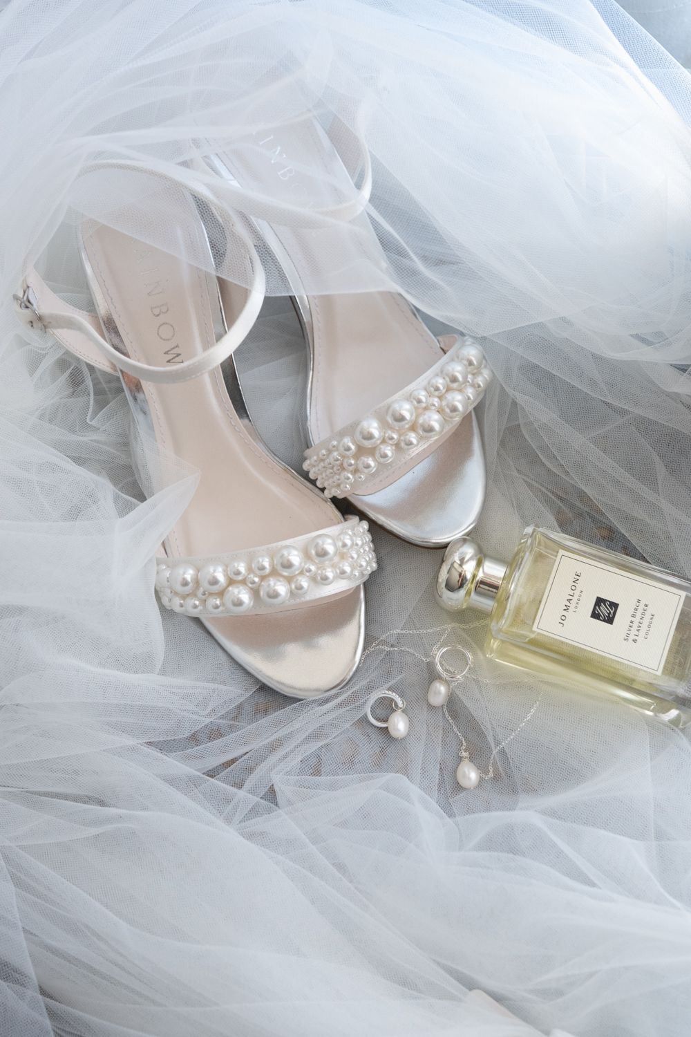 Elegant bridal heels with pearl embellishments rest on delicate white tulle fabric next to a perfume bottle.