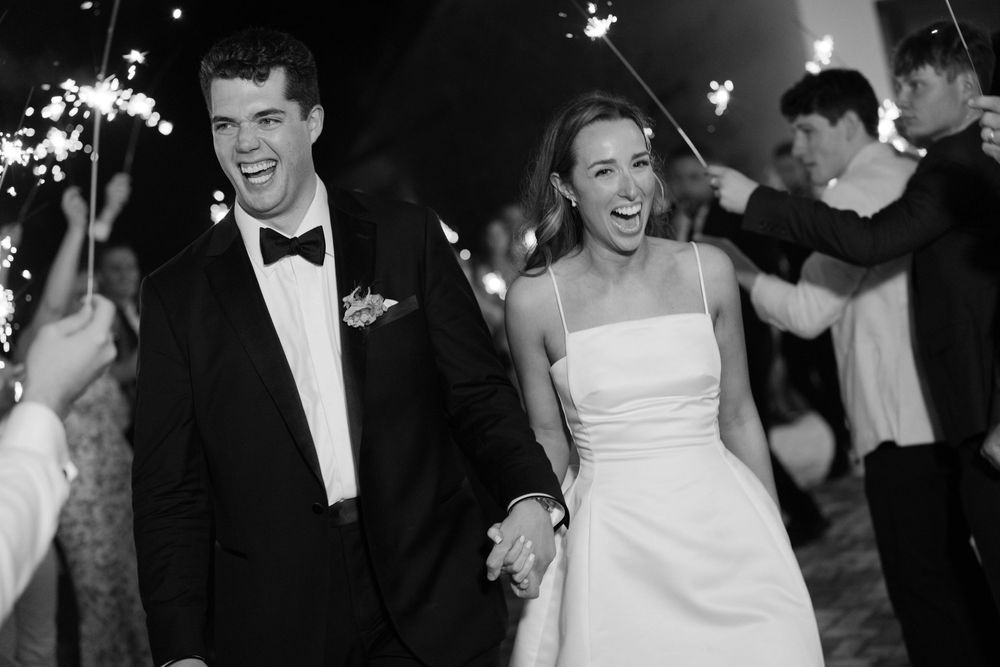 A joyful wedding couple walks through sparklers during their evening reception celebration in black and white photography.