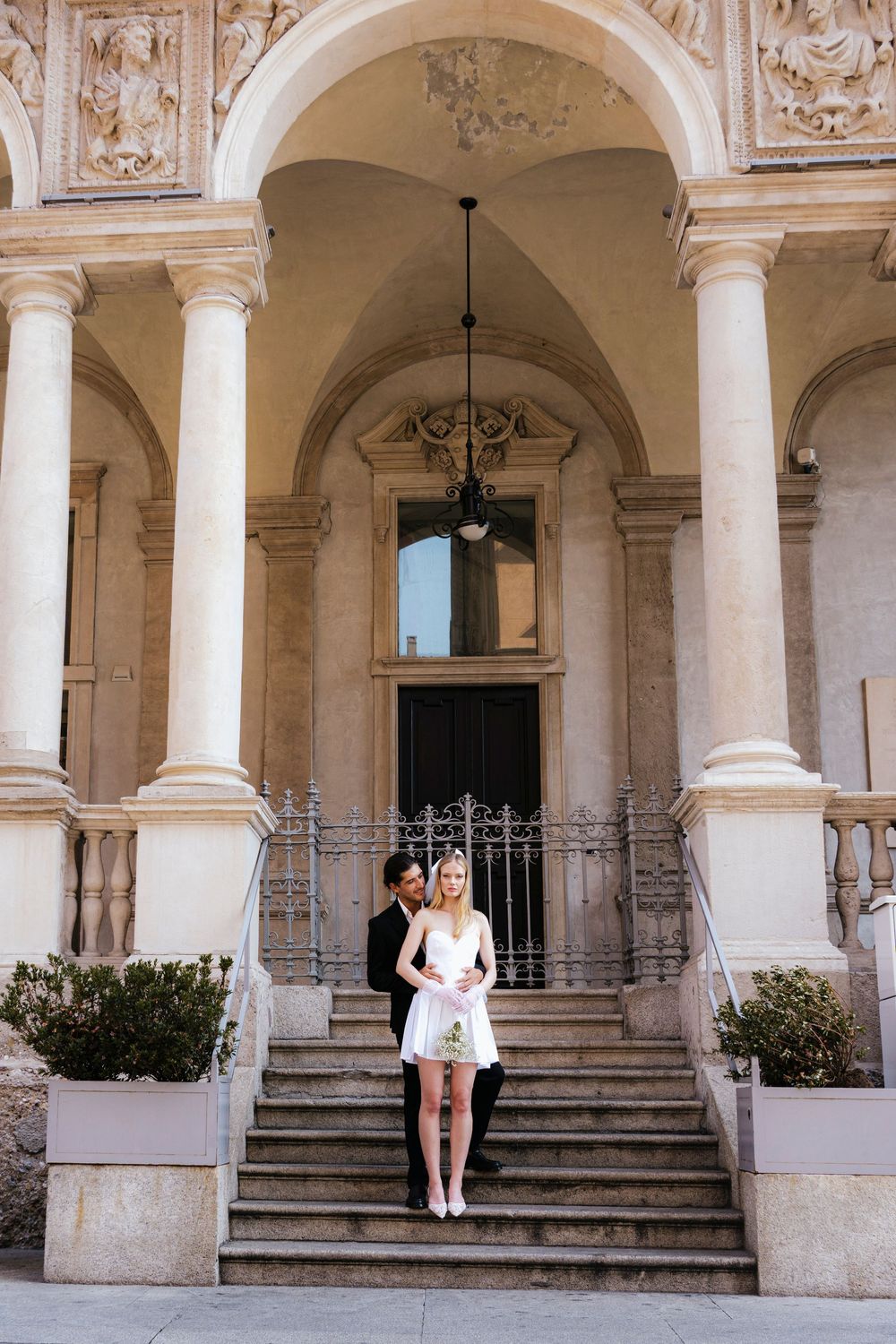 A white dress-clad figure stands on grand stone steps beneath ornate classical archways and columns of a historic building.