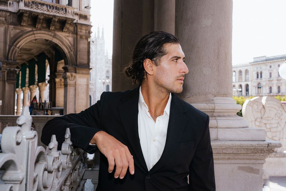 Stylishly dressed figure in black blazer and white shirt leans against ornate stone column at historic European building.