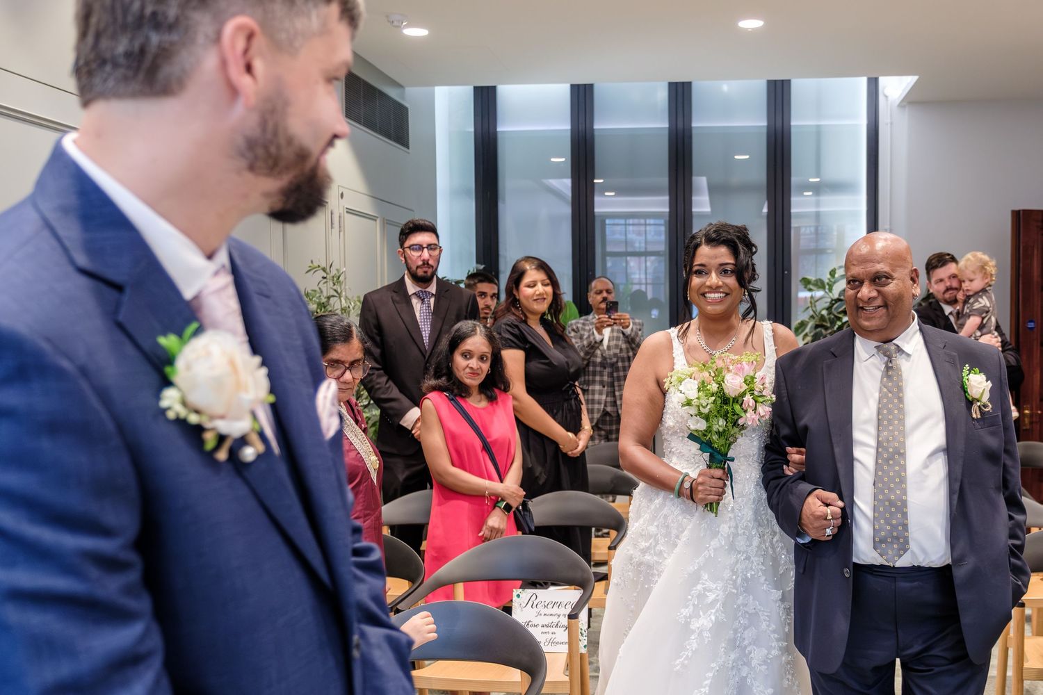 Wedding guests mingling and dancing in a reception hall with large windows and natural lighting.