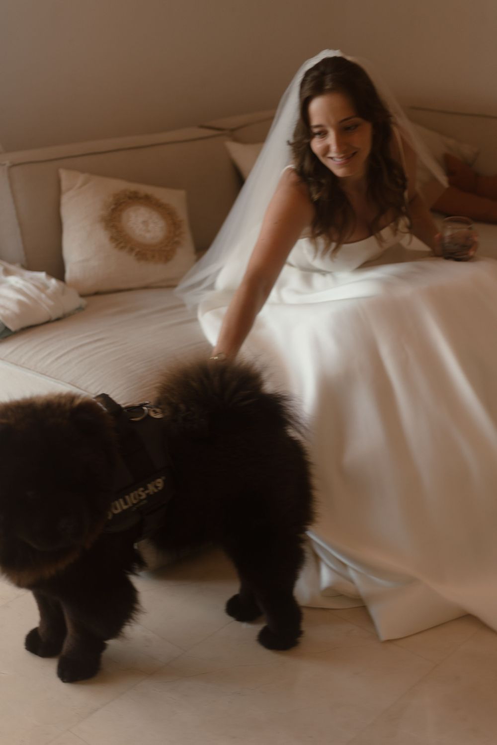 A bride in a white wedding dress sits on a couch petting a fluffy black dog while smiling warmly.