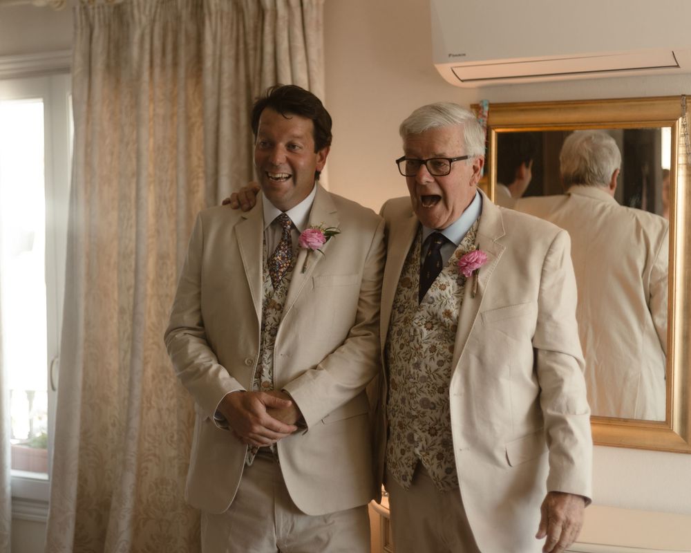 Two people wearing light colored suits and pink boutonnières stand together in a formal setting.