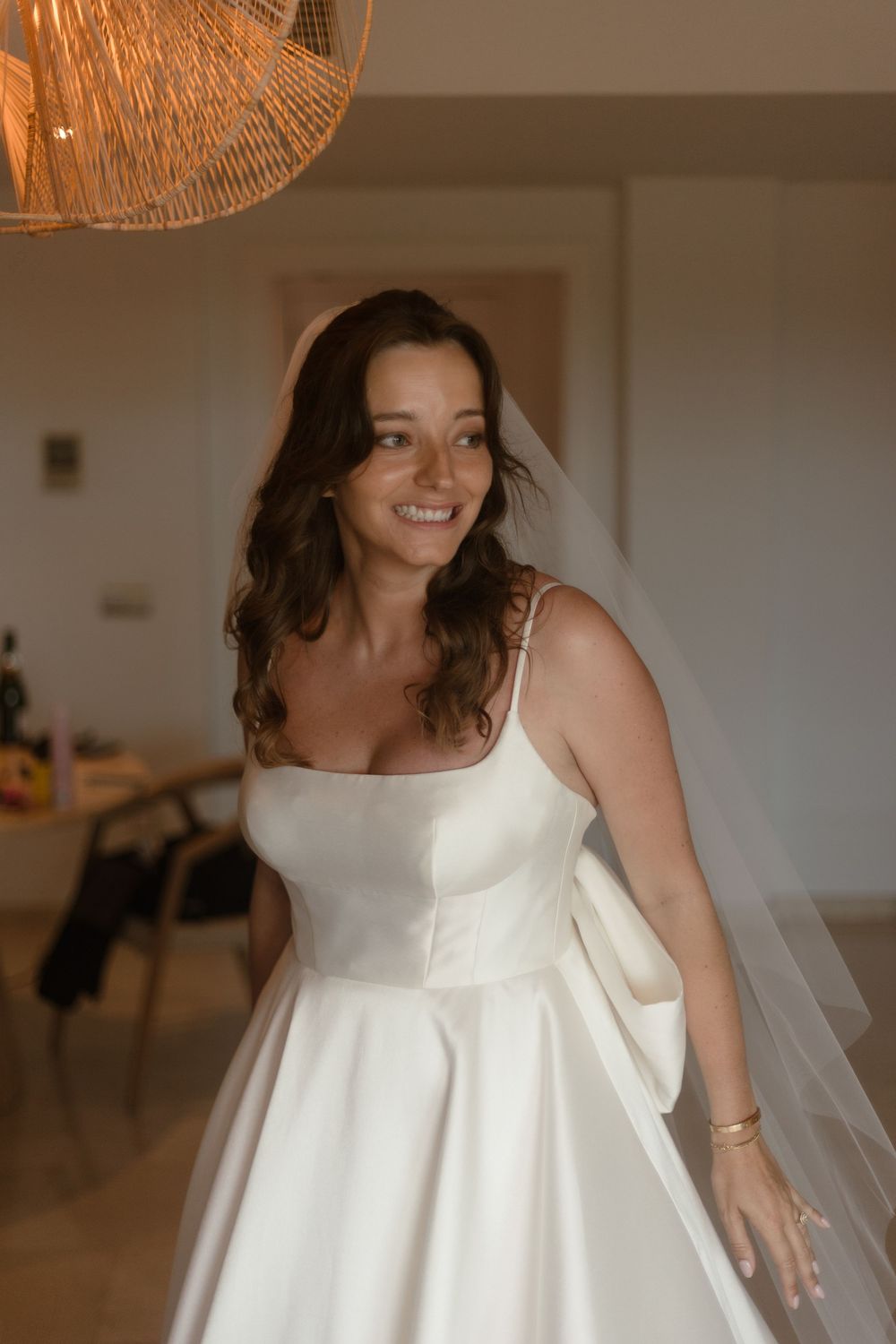 A person wearing a classic white wedding dress with a structured bodice and full skirt smiles indoors under warm lighting.