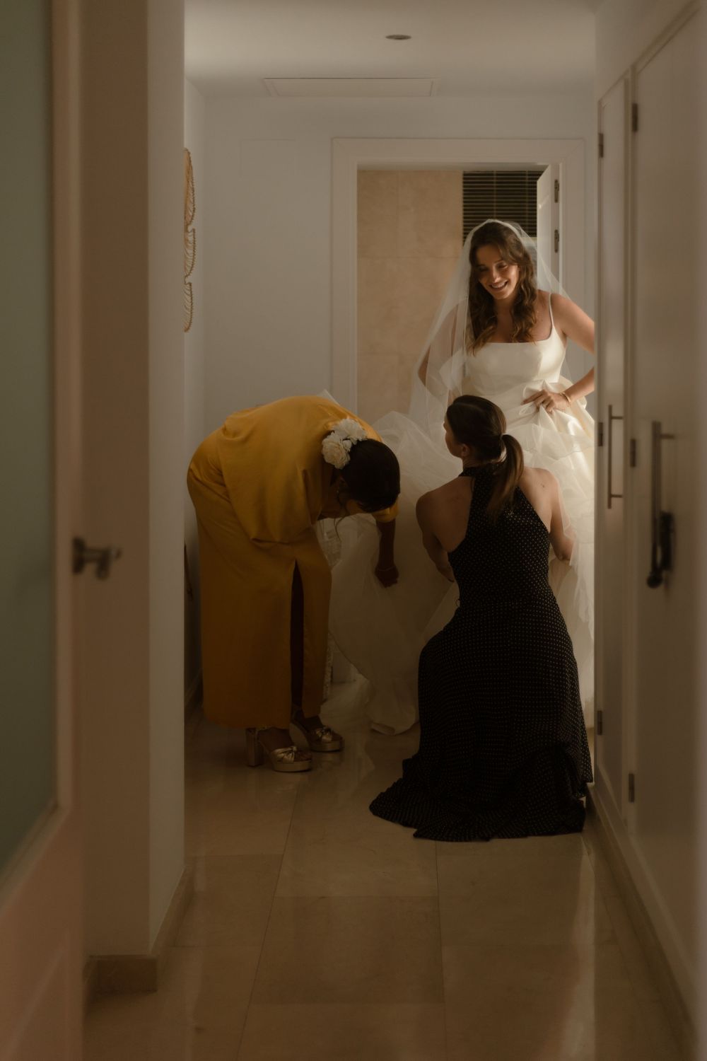 Two people assist with adjusting a white wedding dress in a dimly lit hallway.