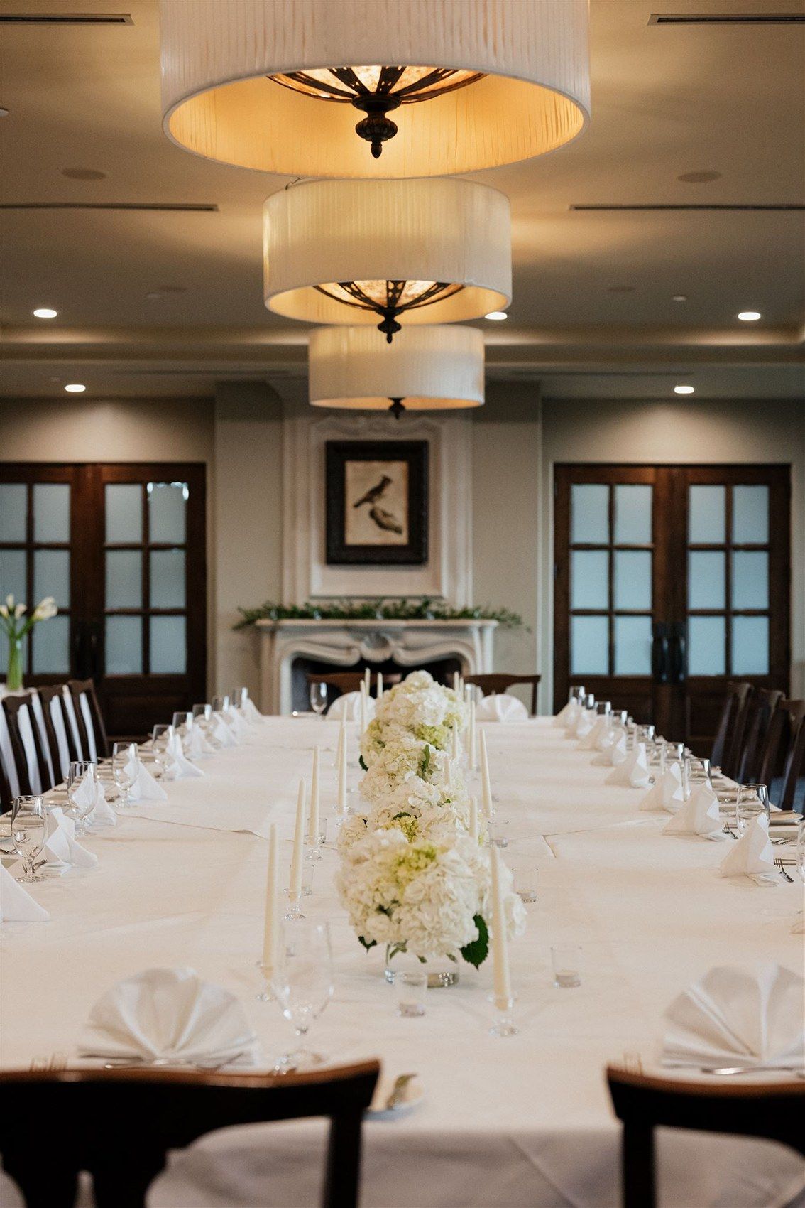Elegant dining table set with white linens and floral centerpiece under tiered pendant lights in an upscale restaurant setting.