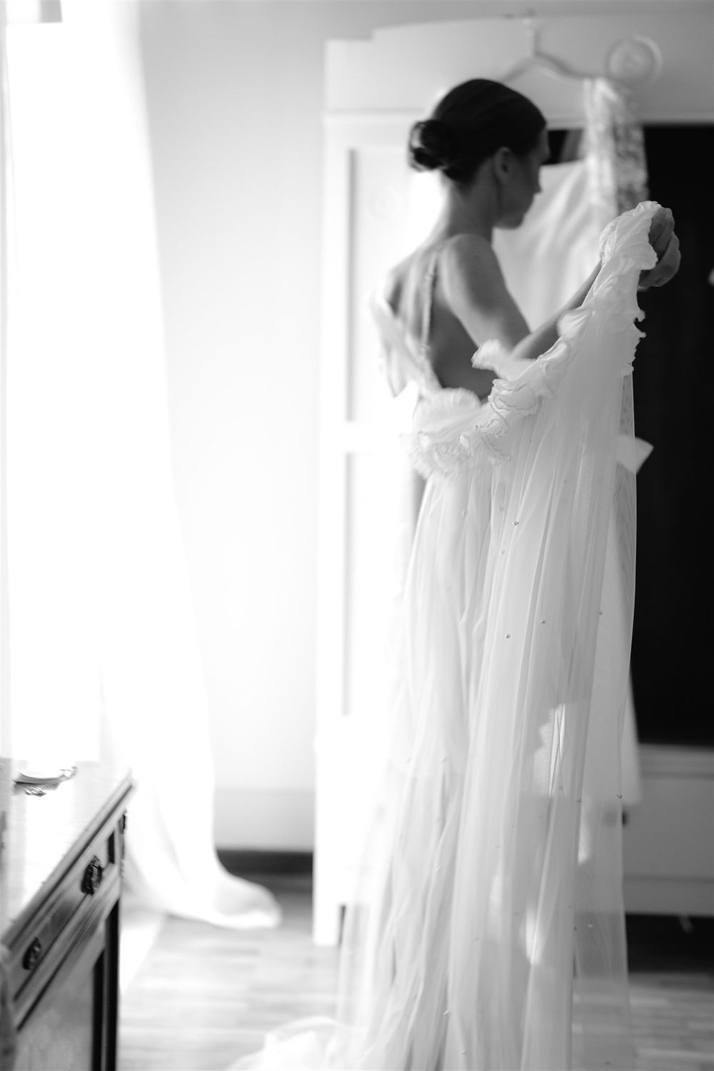 Black and white photo of a wedding dress with delicate lace details being prepared in a sunlit room.