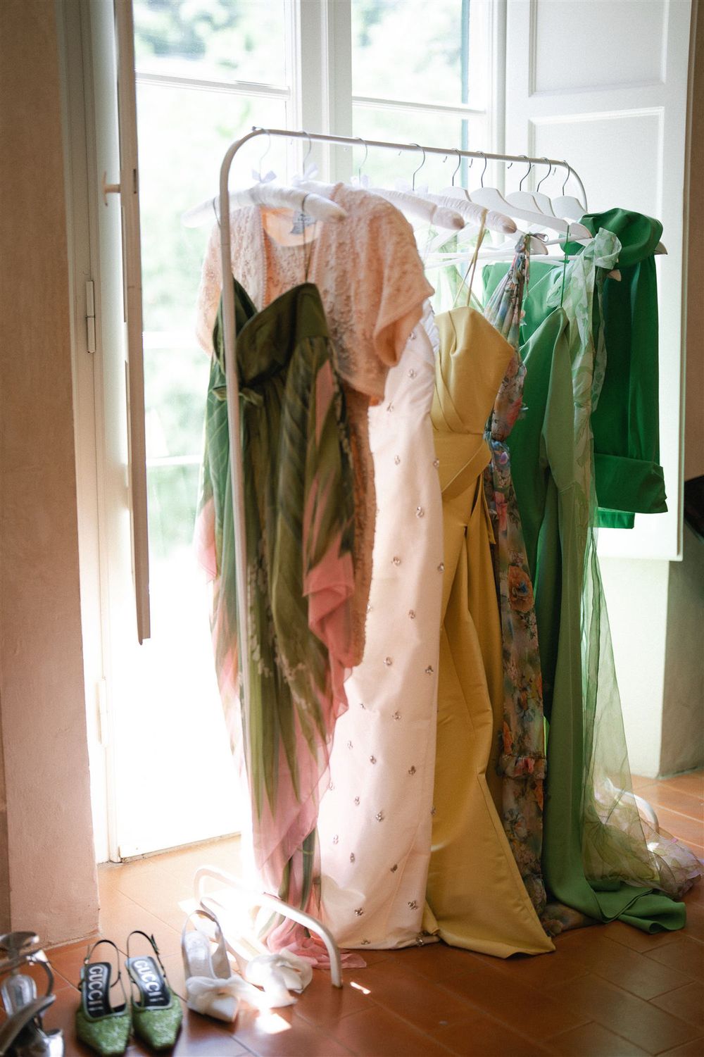 Colorful bridesmaid dresses in green, pink and yellow hanging on a clothing rack near a window.