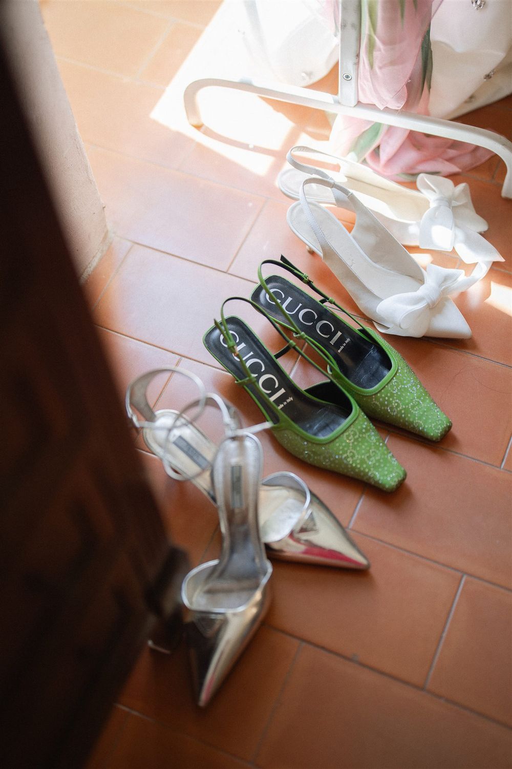 Wedding shoes including sparkly green Gucci flats and silver heels arranged on terracotta floor tiles.