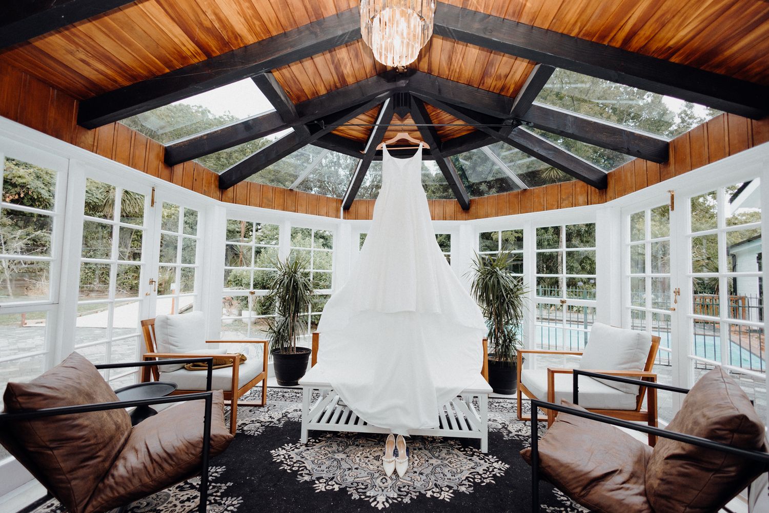 A white wedding dress hangs elegantly in a sunlit conservatory wcaptured by Apertured Media Auckland Wedding Photographer.