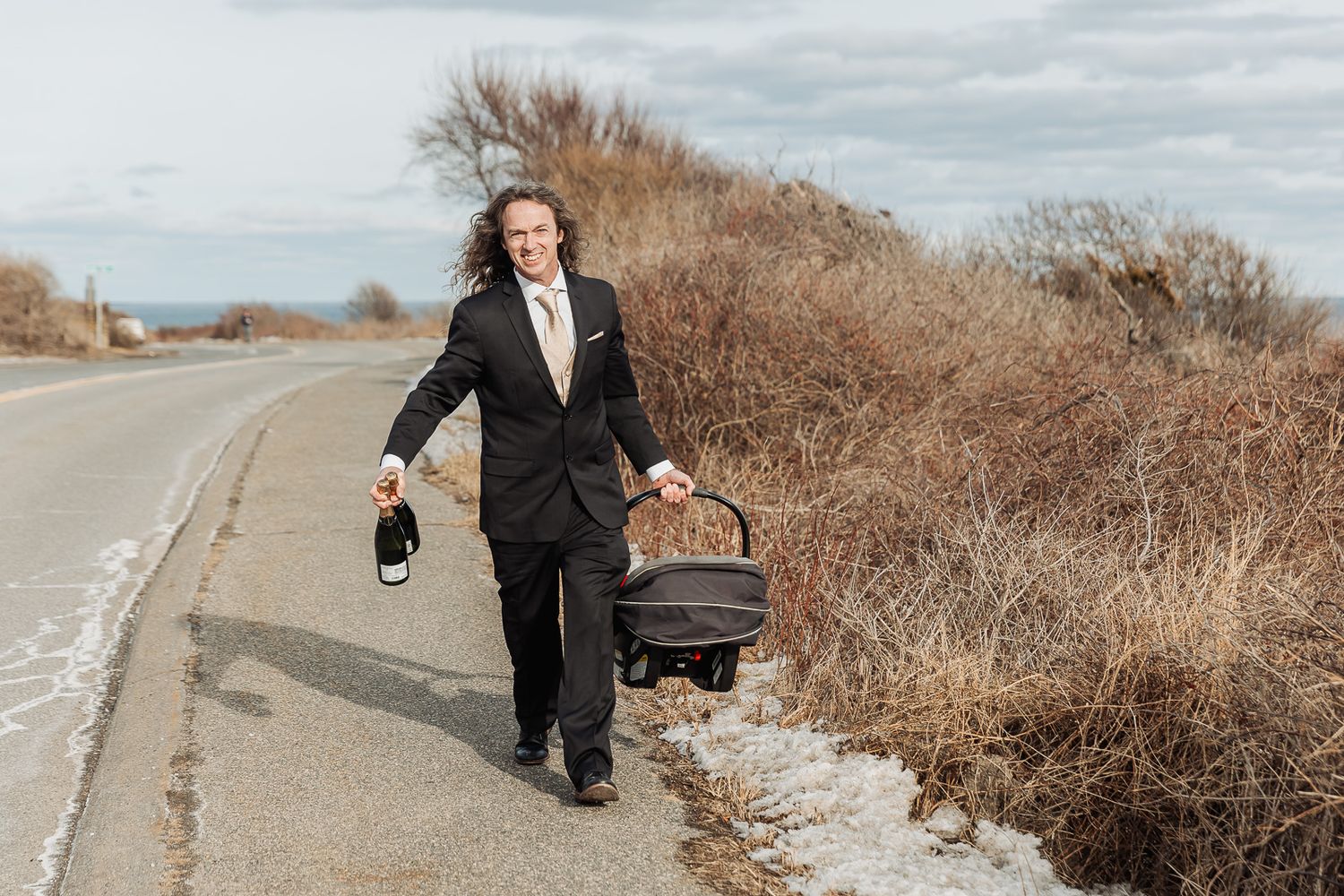 A person in a formal black suit walks along an empty roadside carrying a briefcase and shoes in autumn scenery.