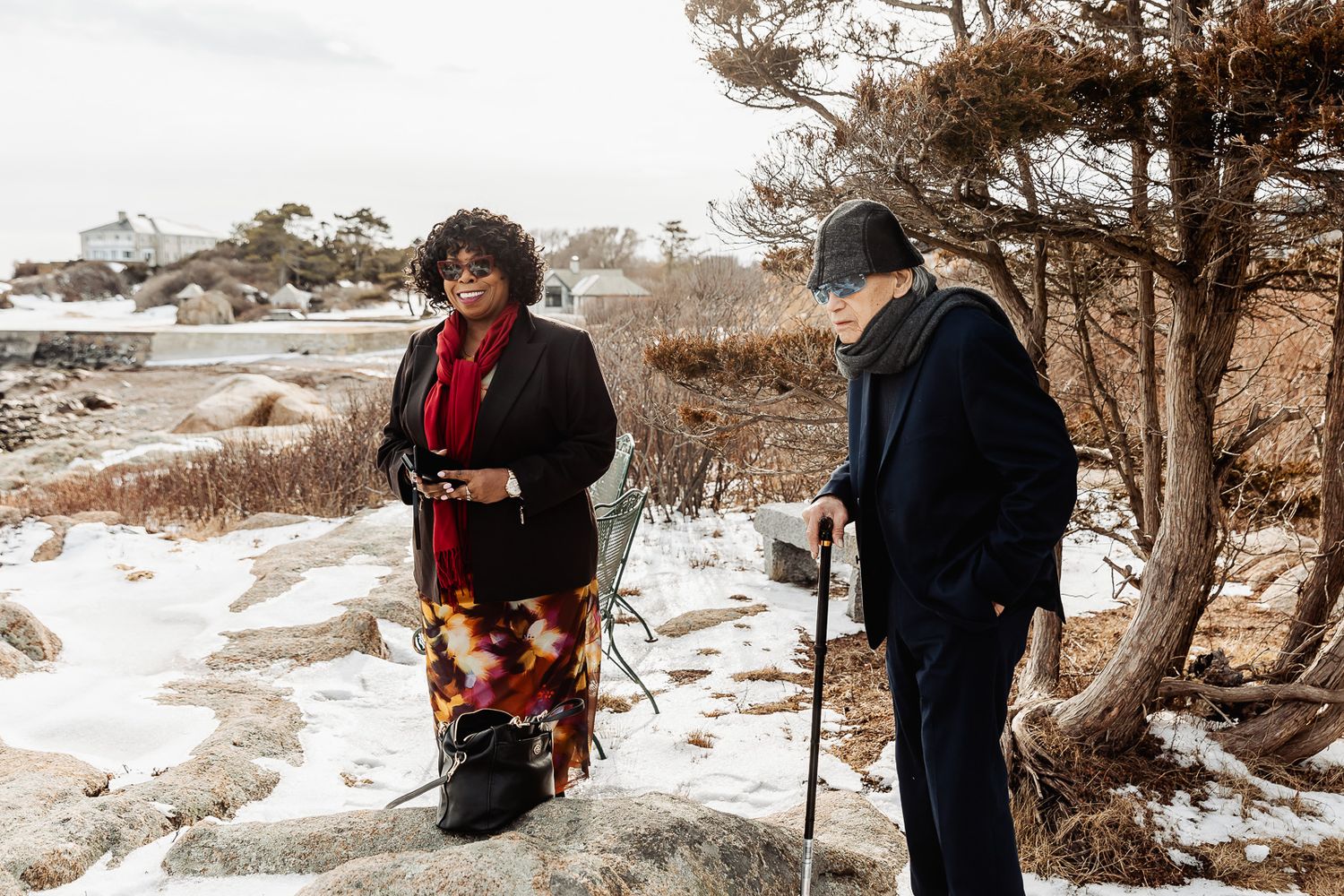 Two people walk with canes along a snowy coastal path bordered by winter vegetation.