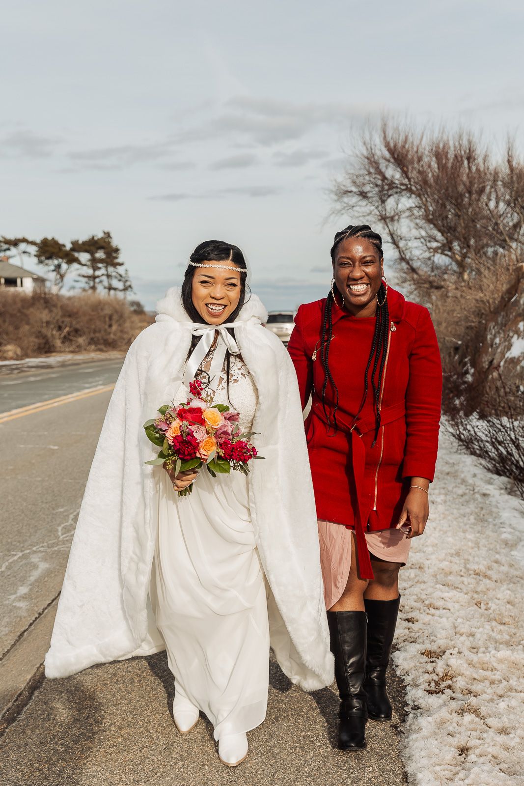 A couple in winter wedding attire poses on a snowy road with evergreen trees in the background.