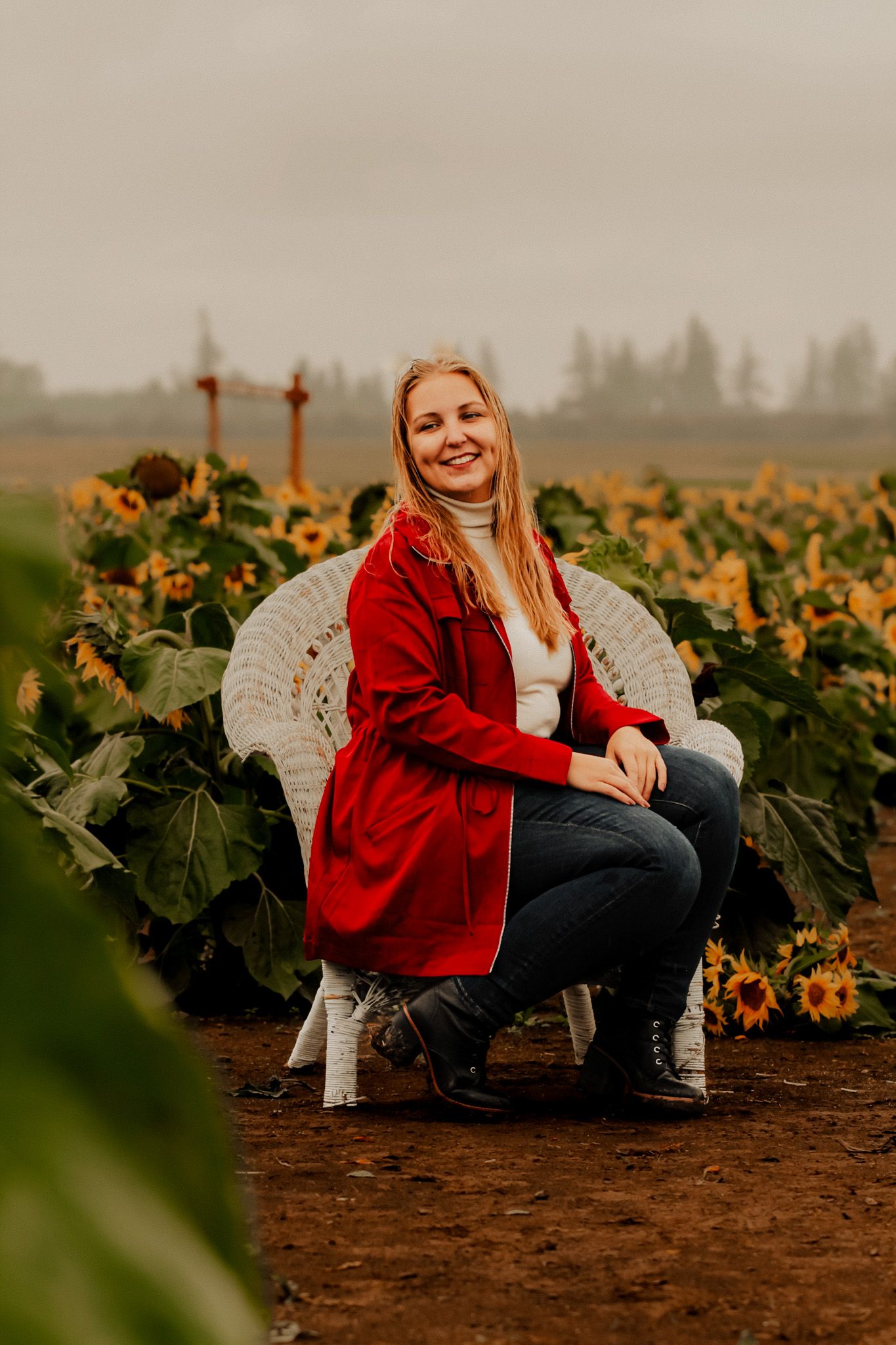 Photographer in red jacket sits among vibrant sunflower field during overcast day, creating rustic autumn atmosphere.