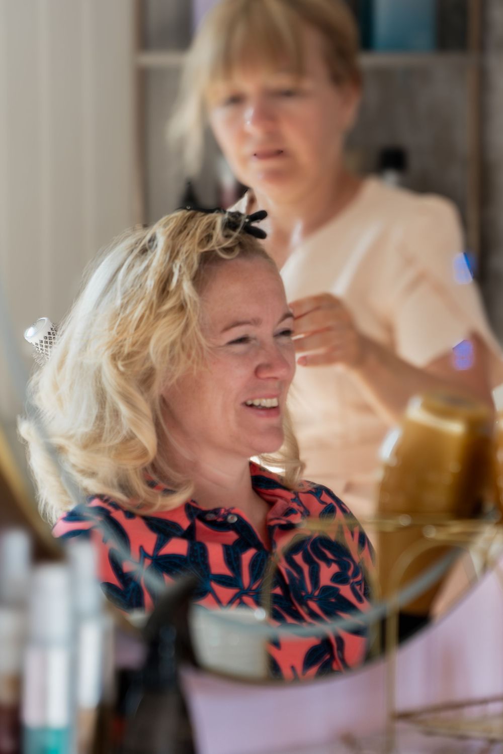 A person in a pink and black patterned top sits while getting their blonde hair styled in a bright indoor setting.