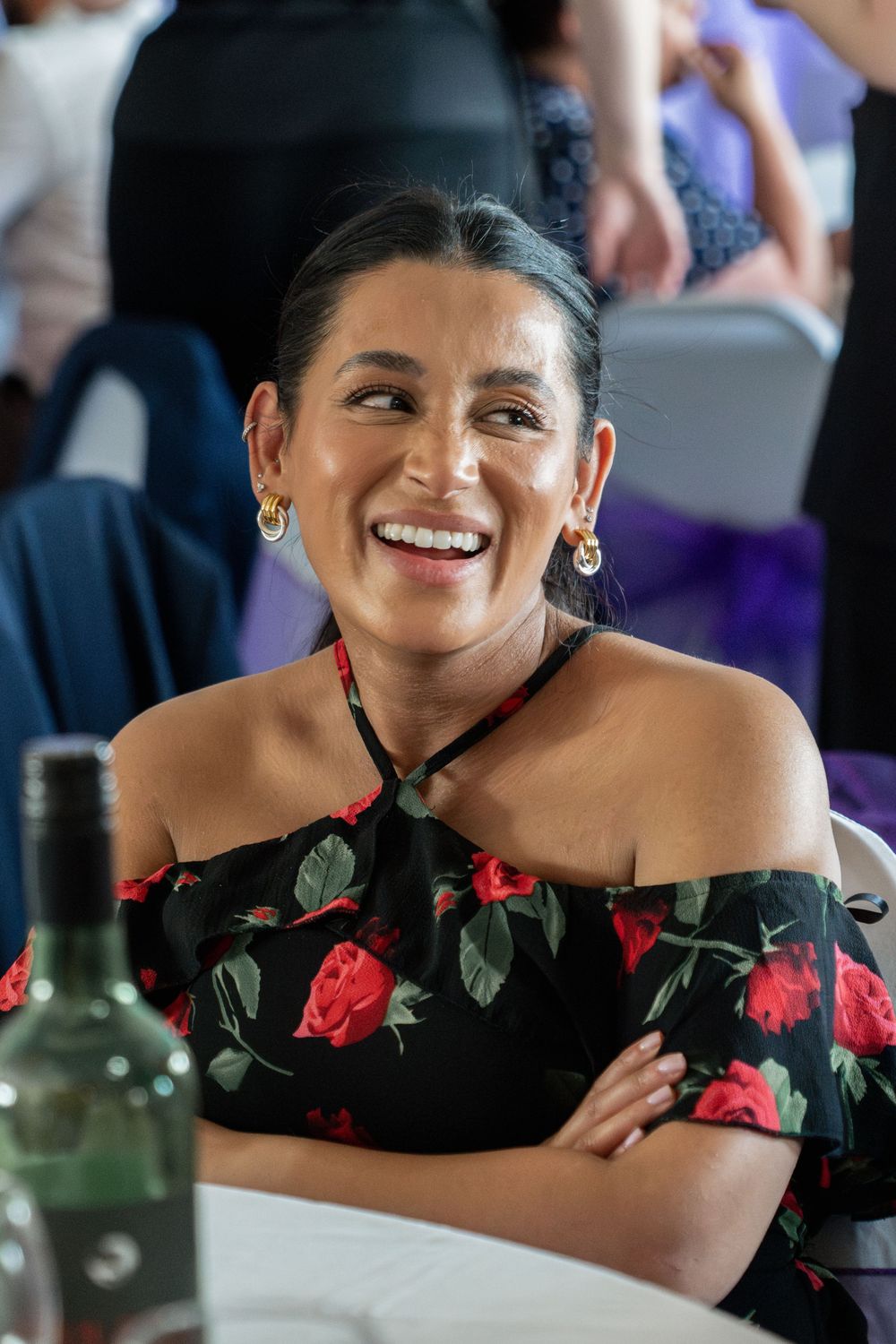 Someone wearing a black floral off-shoulder dress with red roses smiles warmly at a social gathering.