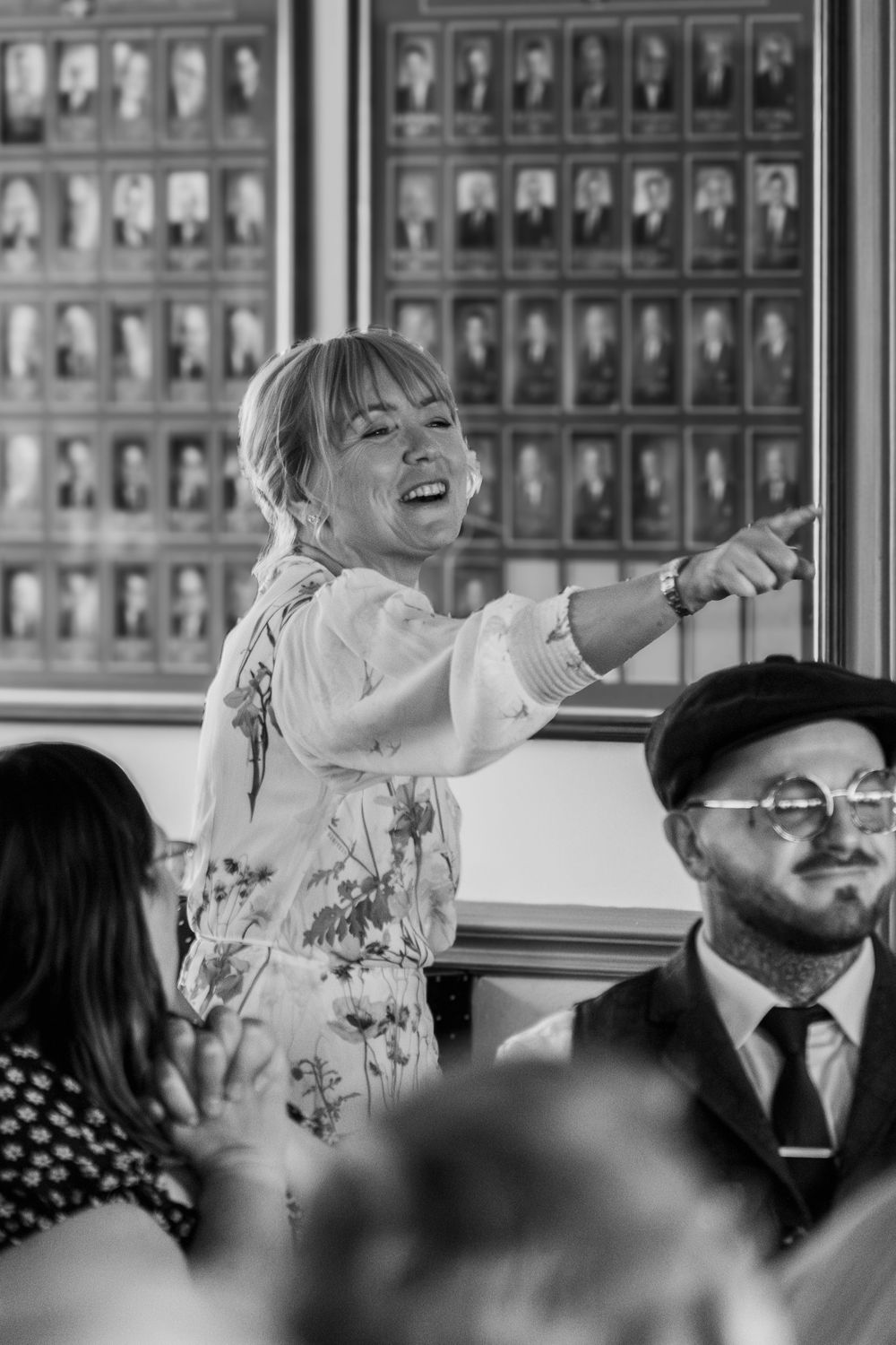 A joyful person in a floral dress laughs while standing in front of a wall displaying black and white portraits.
