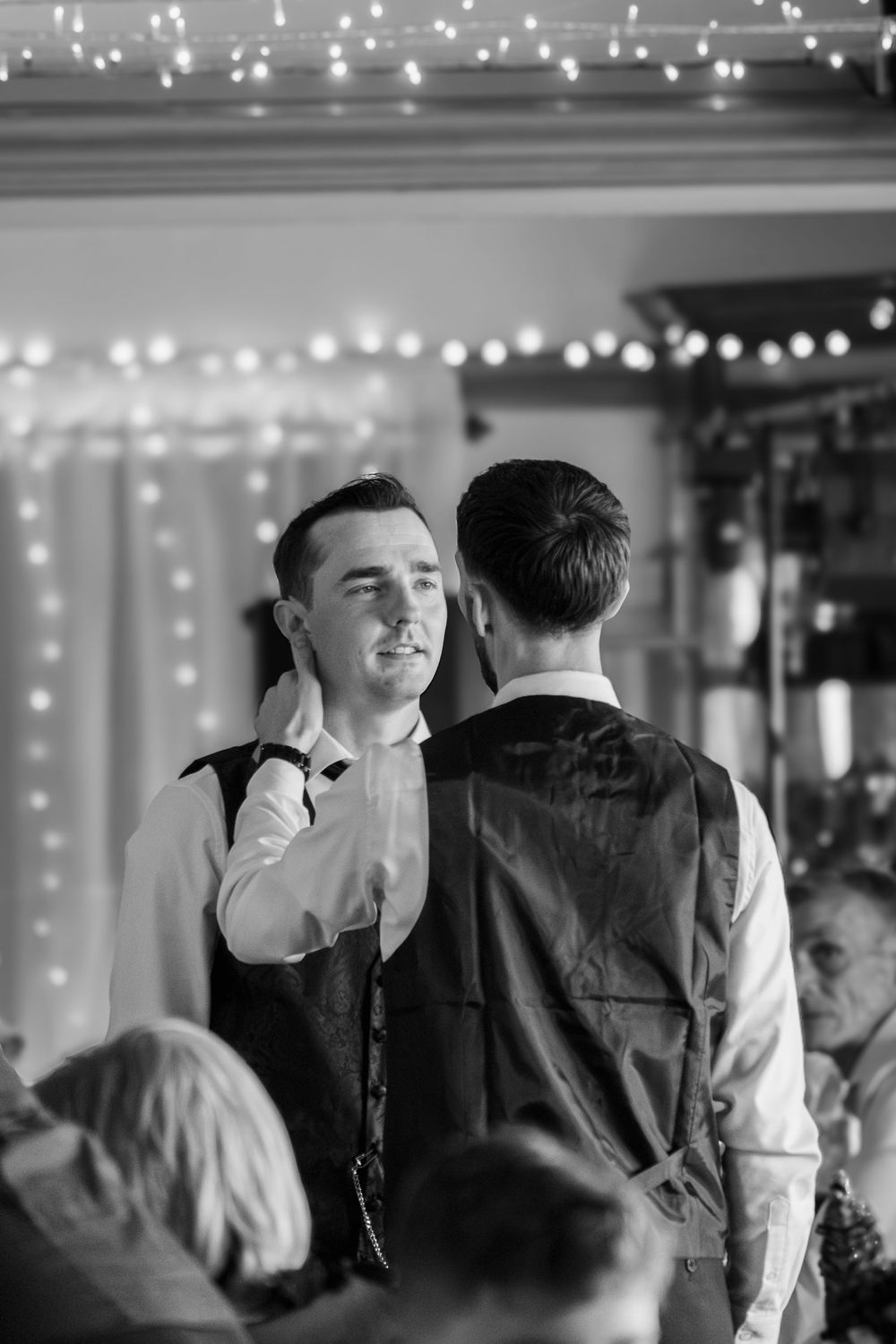 Two figures in formal attire share an intimate moment under string lights at what appears to be a wedding celebration.