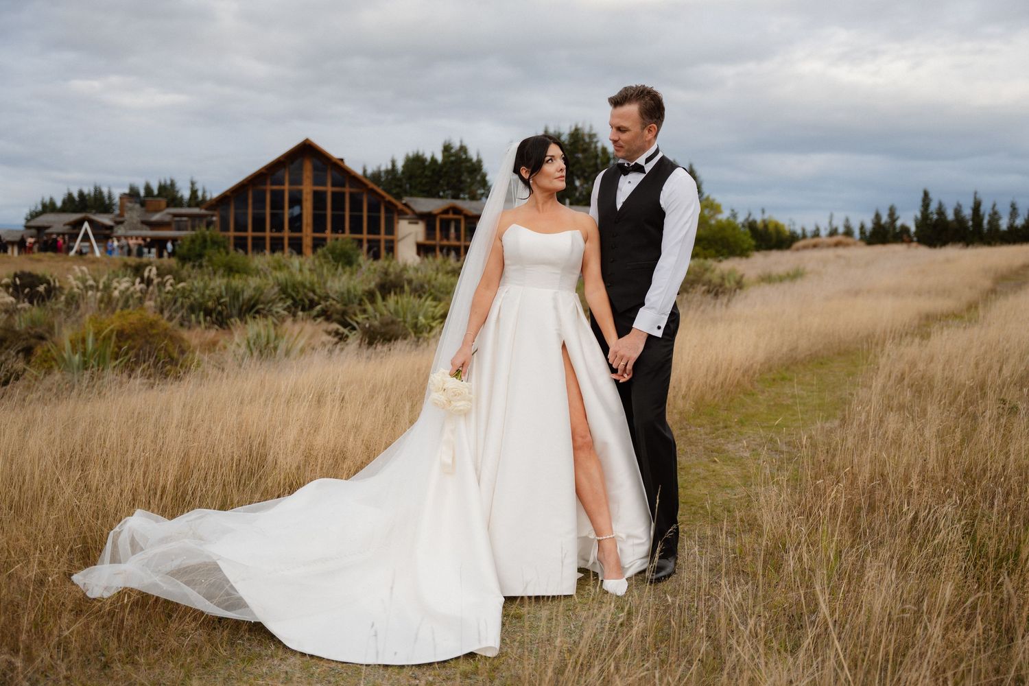 A wedding couple stands in a golden field with a rustic cabin in the background on an overcast day.