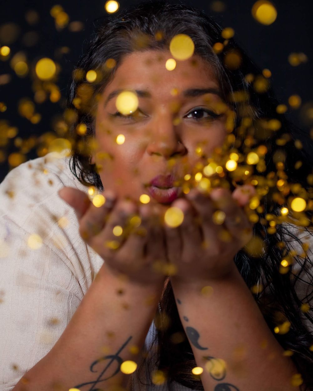 Person in a white outfit blows glitter from their hands against dark background with golden bokeh lights.