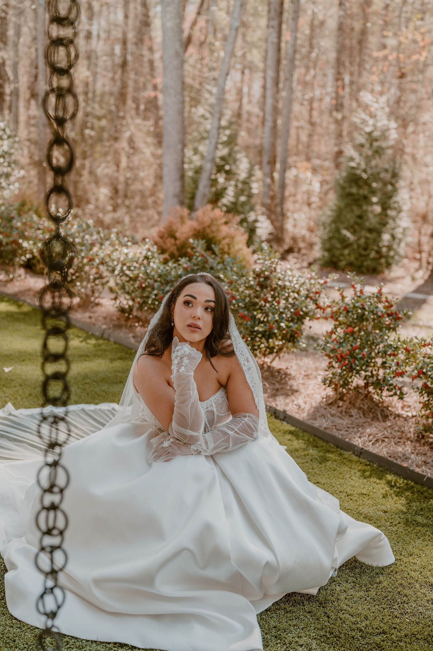A bride in a white flowing gown poses gracefully in an outdoor garden setting with chains in the foreground.