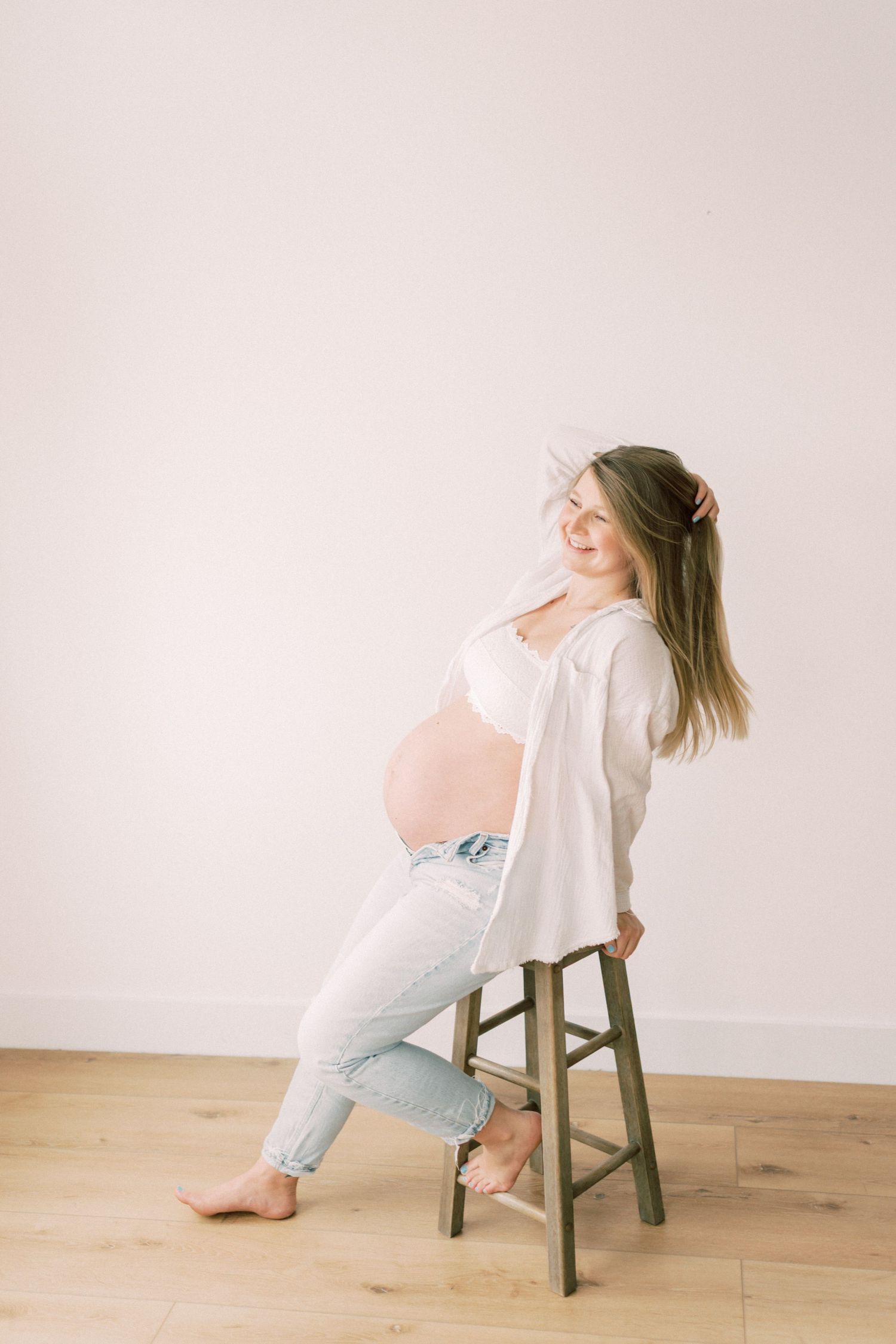 A pregnant woman in casual attire sits on a wooden stool against a pale pink wall in a minimalist setting.