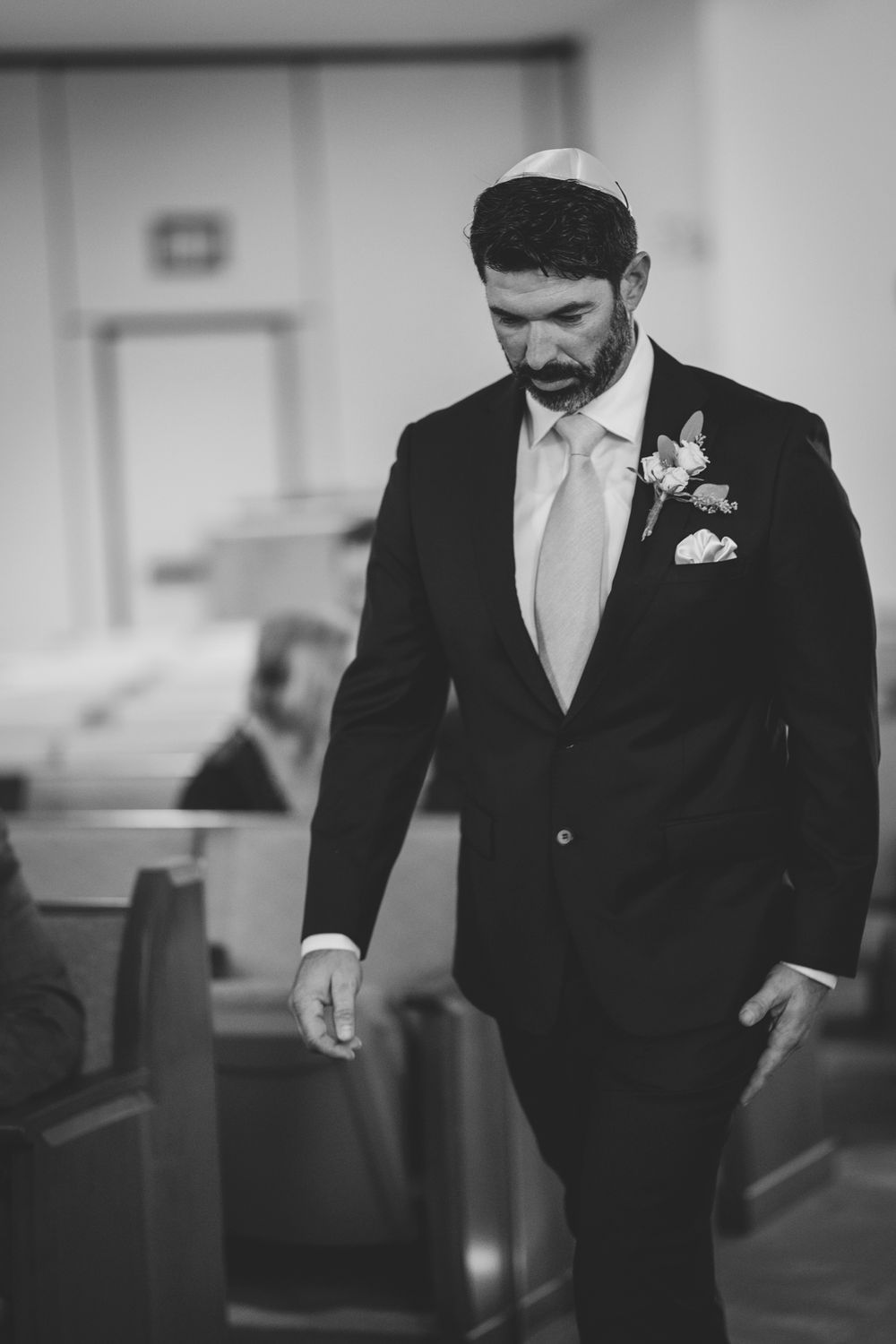 A groom in formal attire with boutonniere walking through church pews.