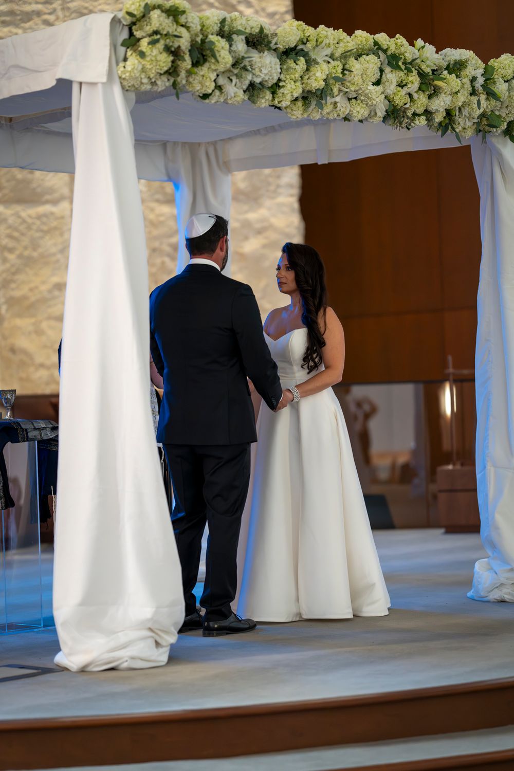 Wedding ceremony under white draped arch decorated with green and white floral arrangements.