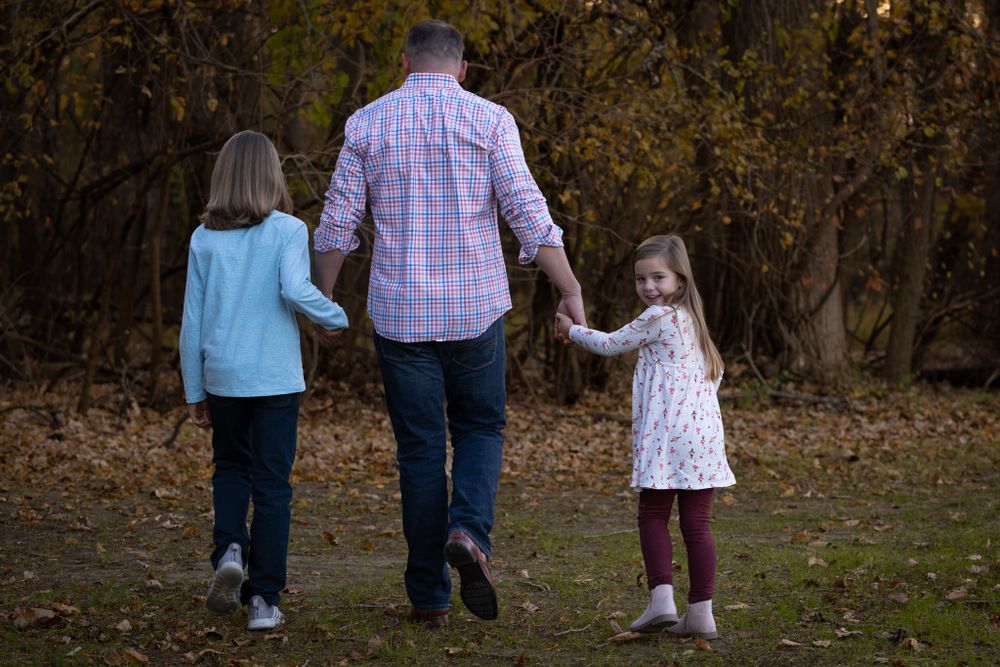 A parent walks hand in hand with two children through a wooded area in fall.