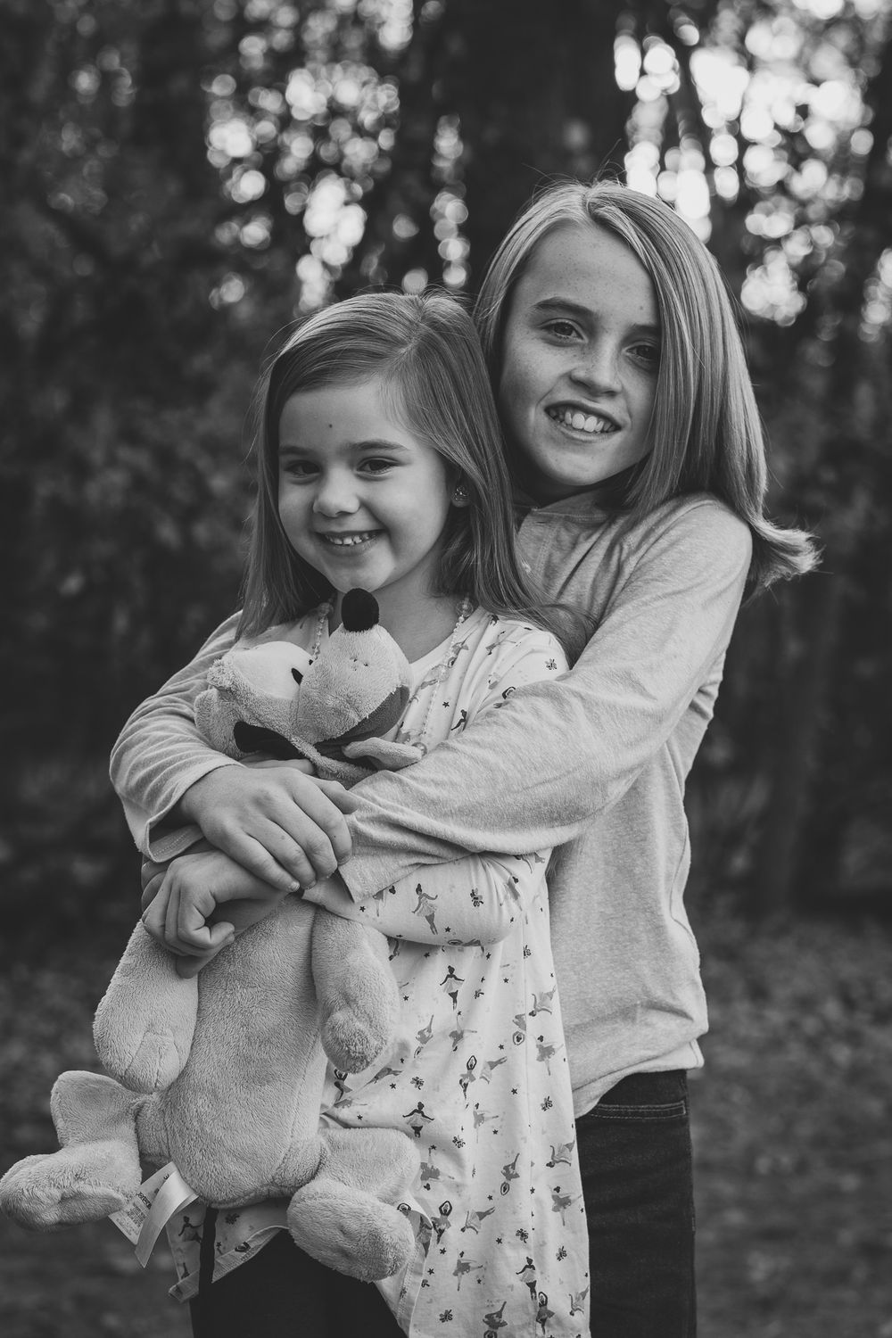 Two people pose together while holding a stuffed animal in a wooded setting.