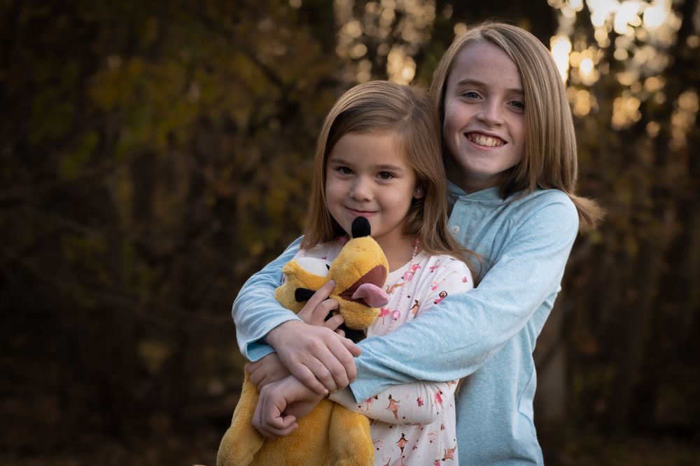 Two children embrace while standing outdoors at sunset in casual attire.