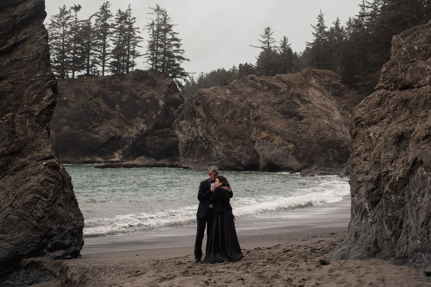 A couple embraces on a moody Pacific Northwest beach surrounded by dramatic rock formations and evergreen trees.