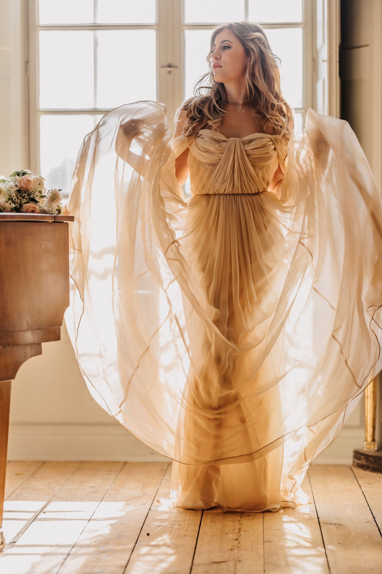Ethereal gold chiffon gown with flowing cape sleeves photographed in sunlit room with wooden floors and floral decor.