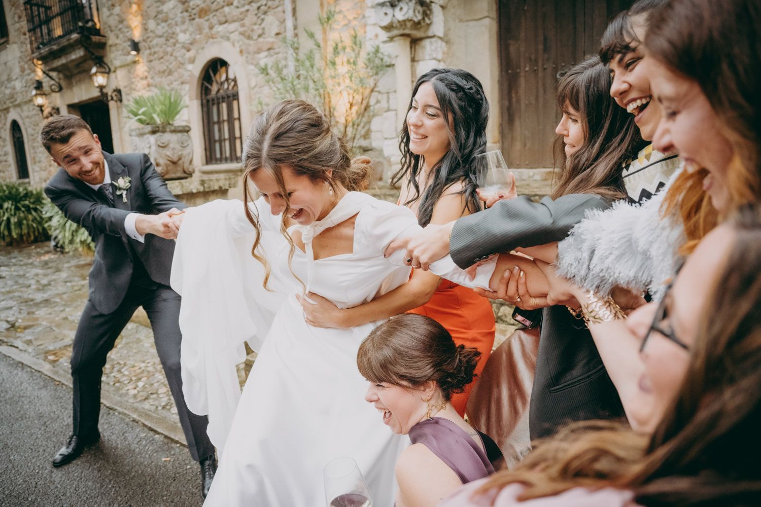Los invitados de la boda tiran juguetonamente del vestido de la novia durante las fotos grupales al aire libre