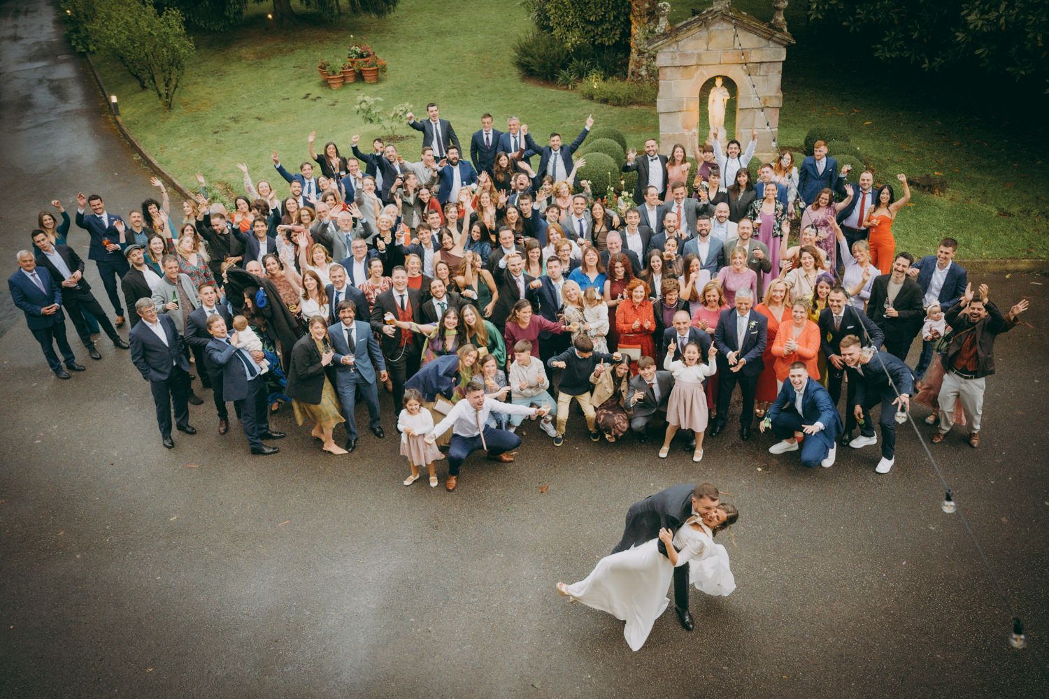 Gran fotografía grupal de boda tomada desde arriba con la pareja acostada en el suelo rodeada de invitados.