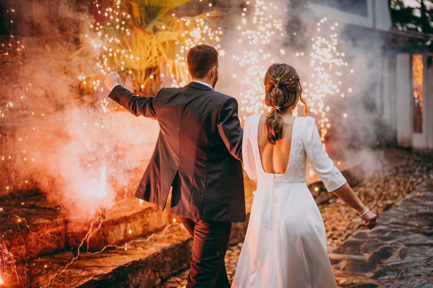 Recién casados tomados de la mano con bengalas creando un efecto dramático en la foto de la boda por la noche.