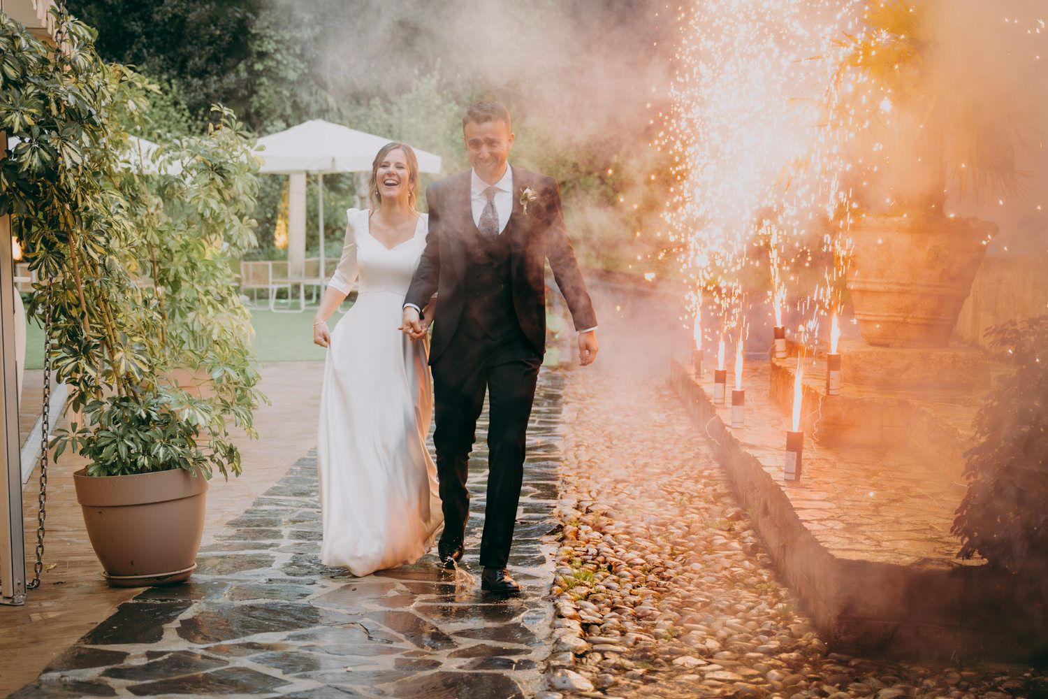Unos novios camina por un camino de piedra con una cálida luz dorada y efectos de humo creando una atmósfera de boda mágica al aire libre.