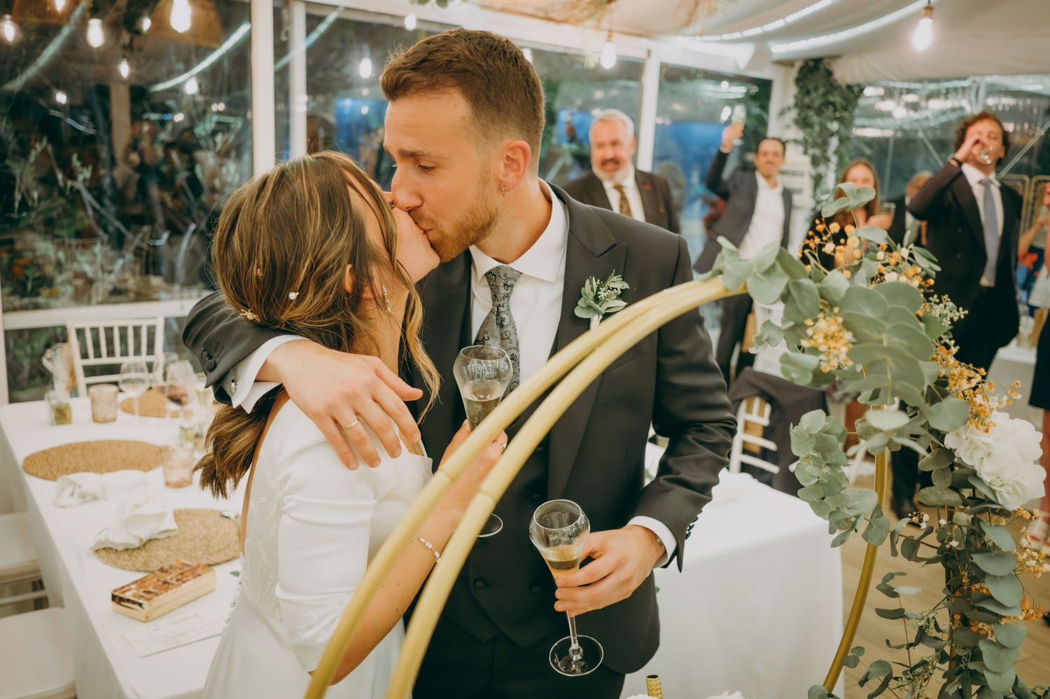 Una pareja de recién casados comparte un tierno beso en la recepción de su boda mientras el novio sostiene una copa de champán.