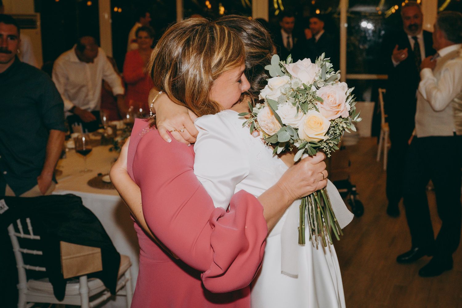 Madre e hija se abrazan en la recepción de una boda mientras una sostiene un ramo de flores blancas mientras los invitados miran al fondo.