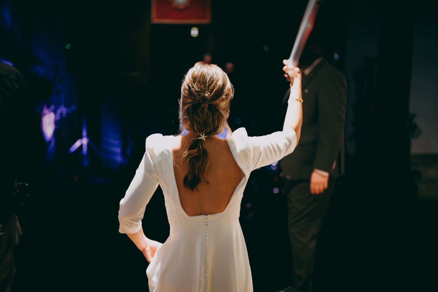 Una novia con un vestido blanco sin espalda levanta un brazo mientras baila en un lugar poco iluminado en el Palacio de Villabona.