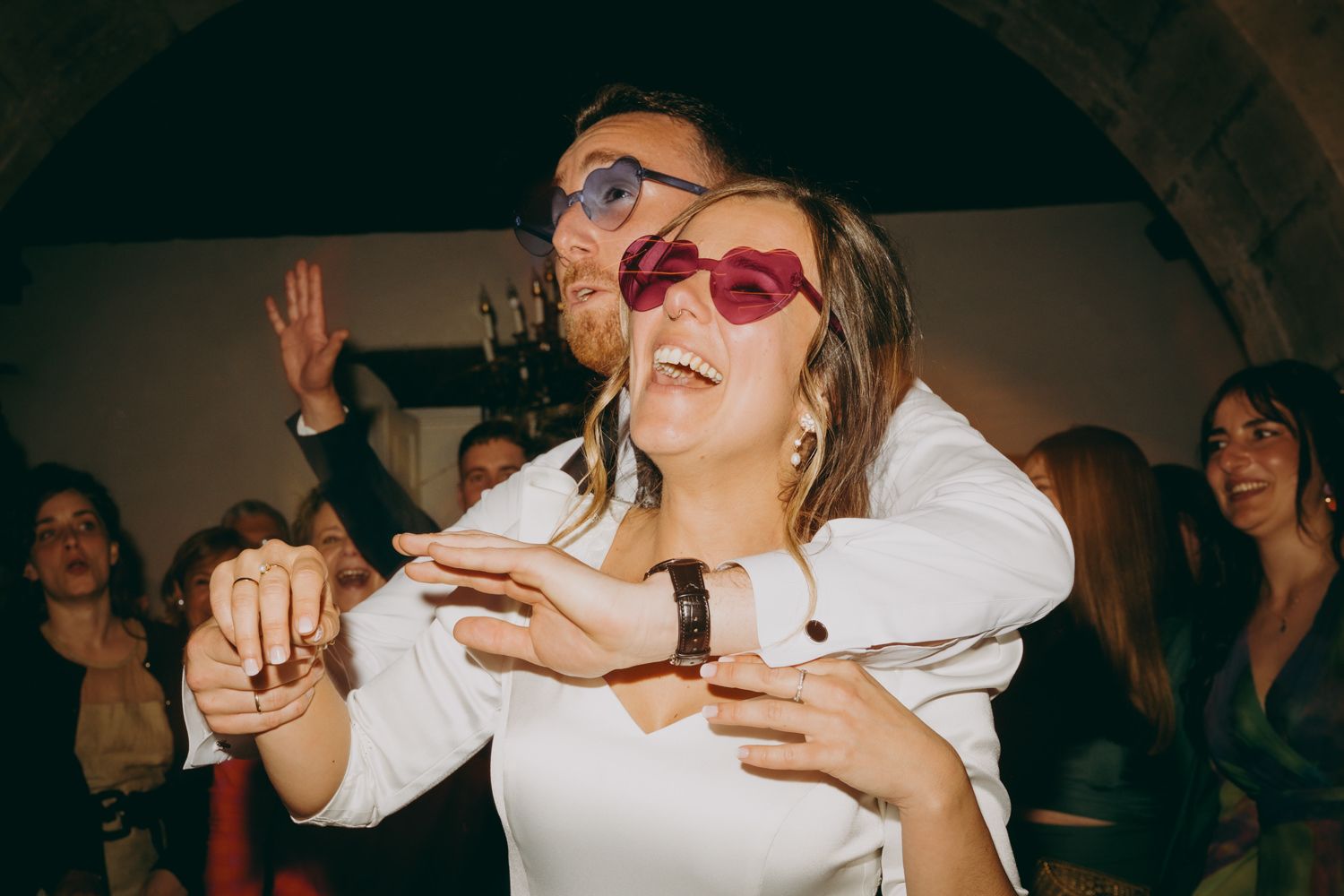 Unos novios con gafas de sol bailan con entusiasmo en una boda con otros invitados visibles en el fondo.